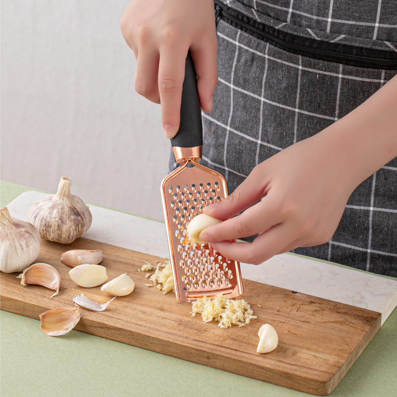 Person using a kitchen household peeler gadget with copper plating to grate garlic on a cutting board.